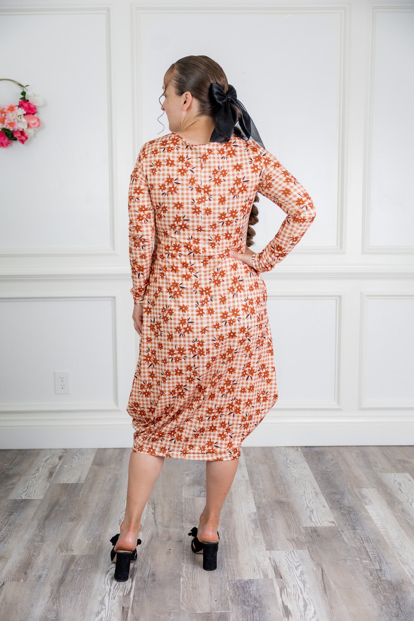 Woman wearing a patterned dress standing in a room with wooden flooring and white paneled walls.