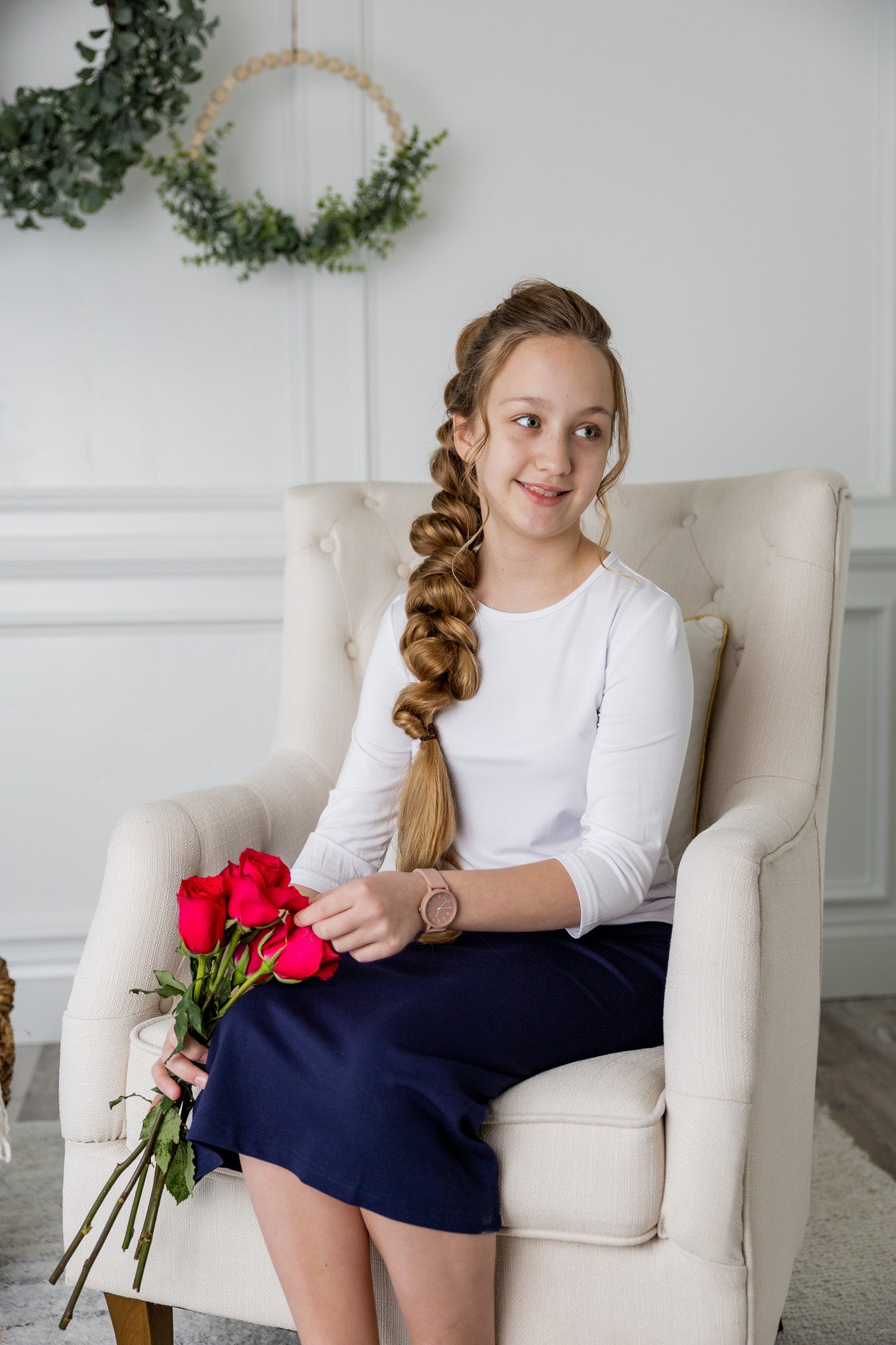 Young girl with braided hair sitting in a chair holding red flowers, wearing a white shirt and navy skirt.