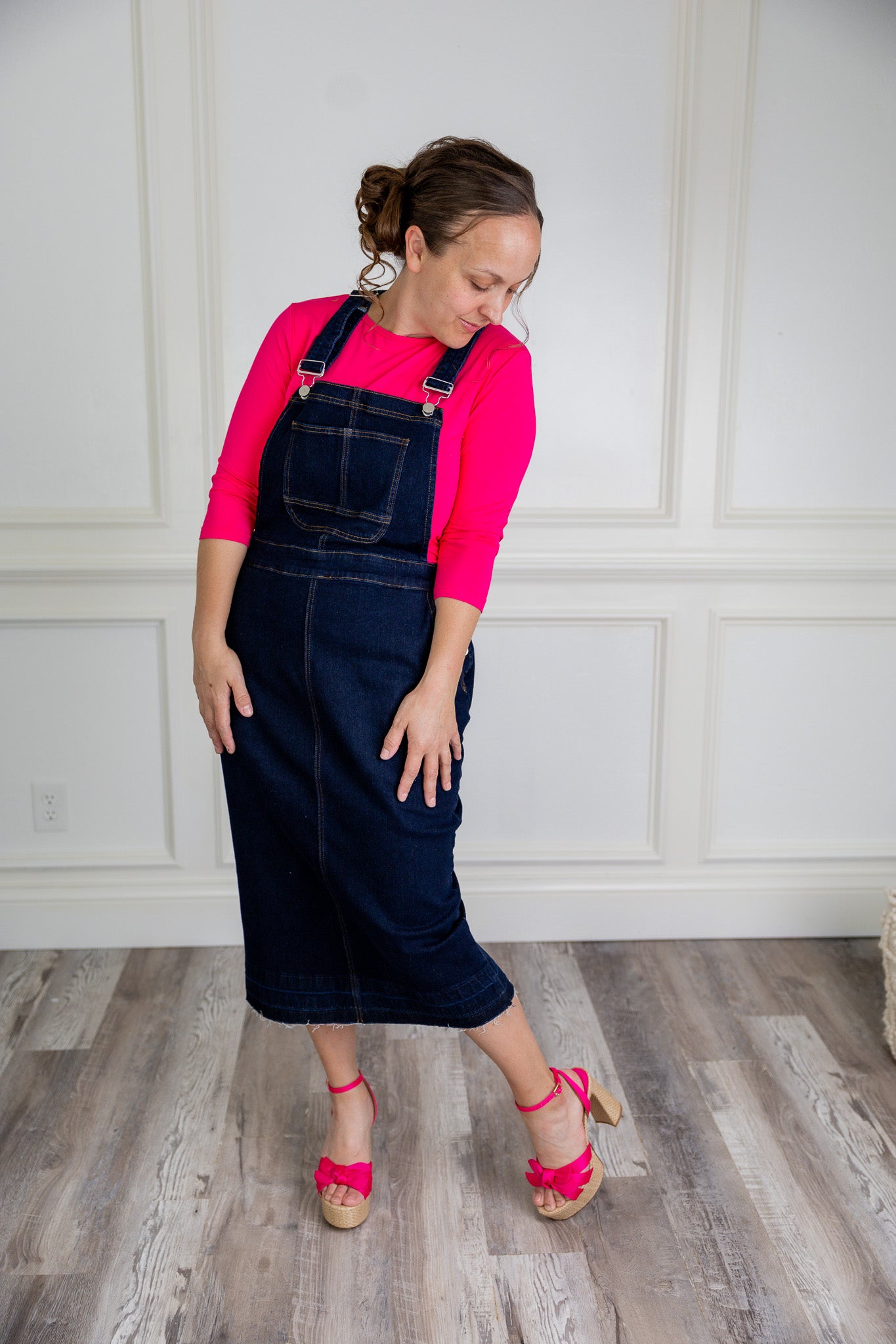 Model wearing a 'NUGGLES® modest dark wash denim overall dress with front pocket and raw hem.