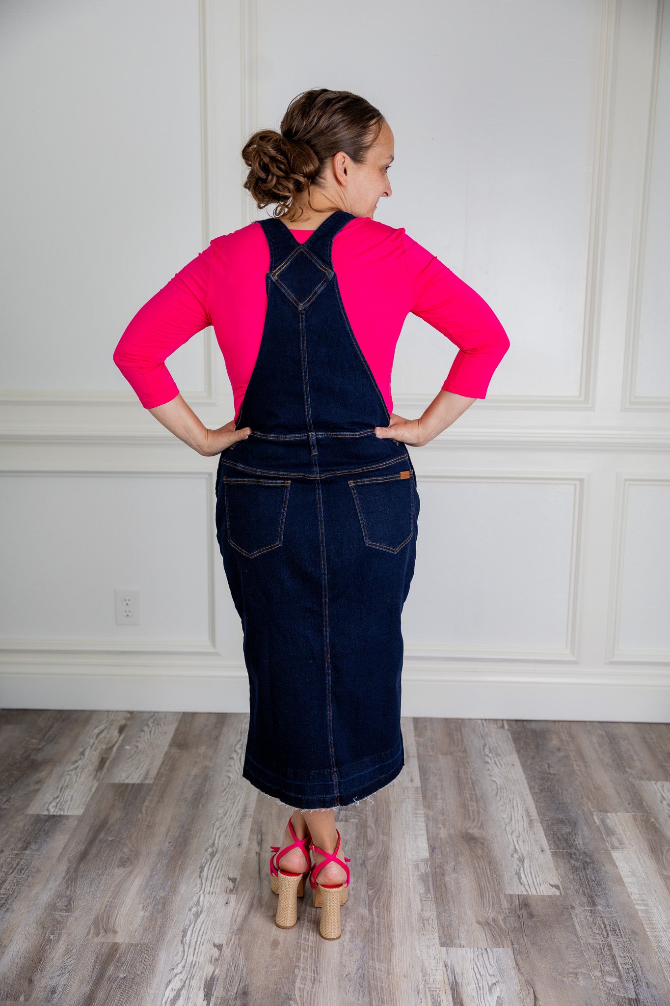 Model wearing a 'NUGGLES® modest dark wash denim overall dress with front pocket and raw hem.