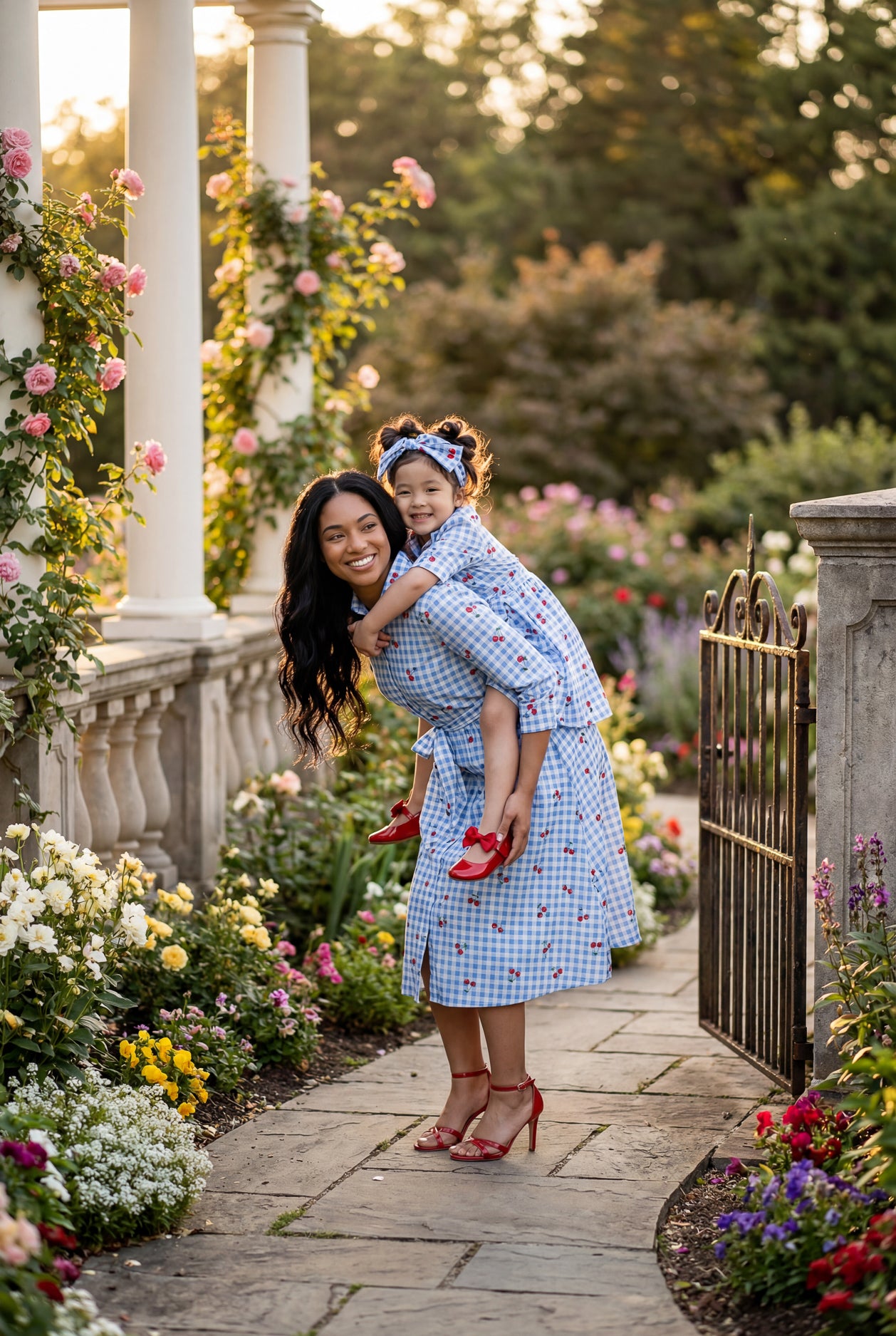 Woman and child in matching dresses standing in a garden with flowers and a gate.