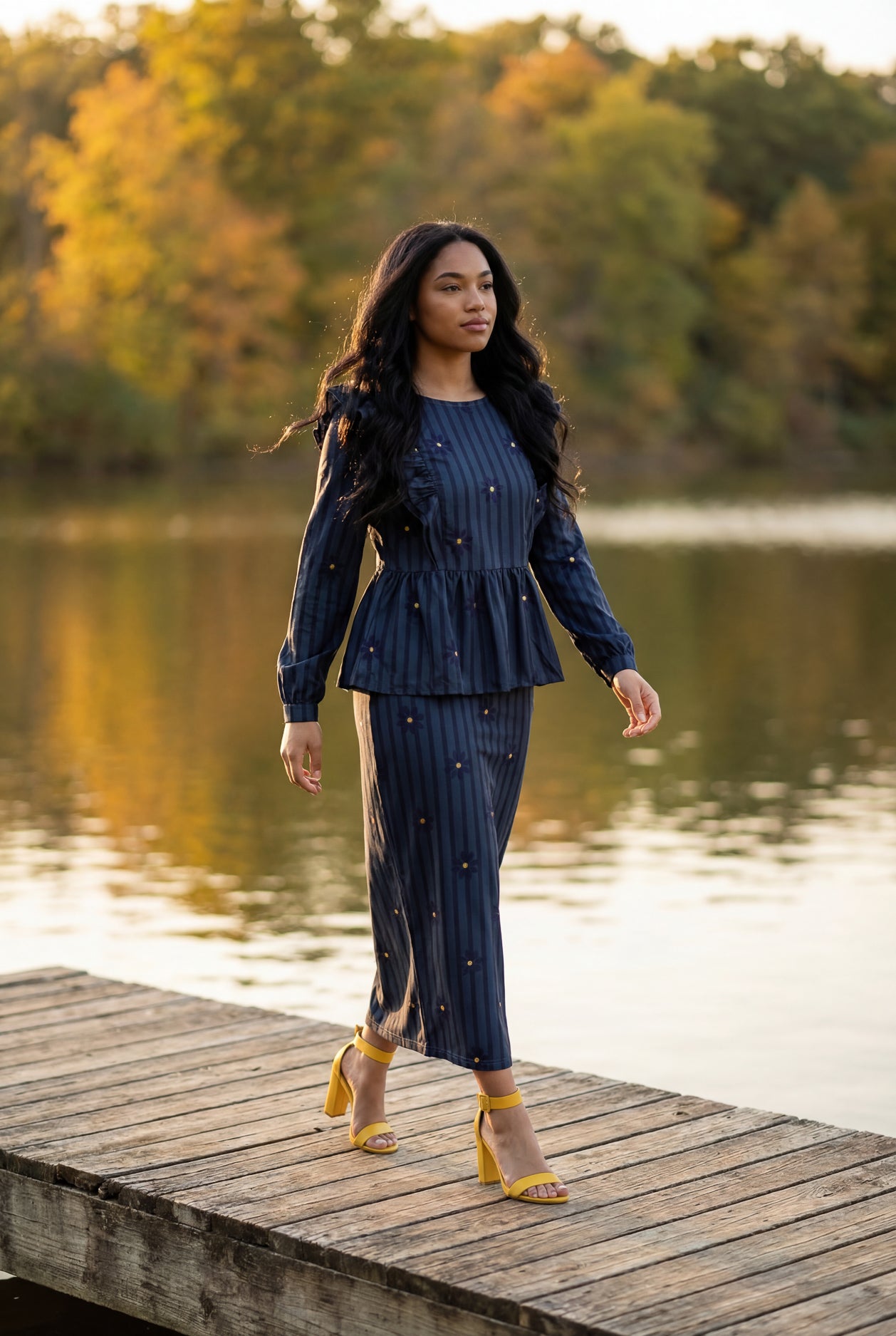 Woman in a navy blue outfit standing on a wooden dock by a lake with trees in the background.