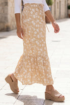 Woman in a white top and yellow floral skirt standing on a stone-paved street.