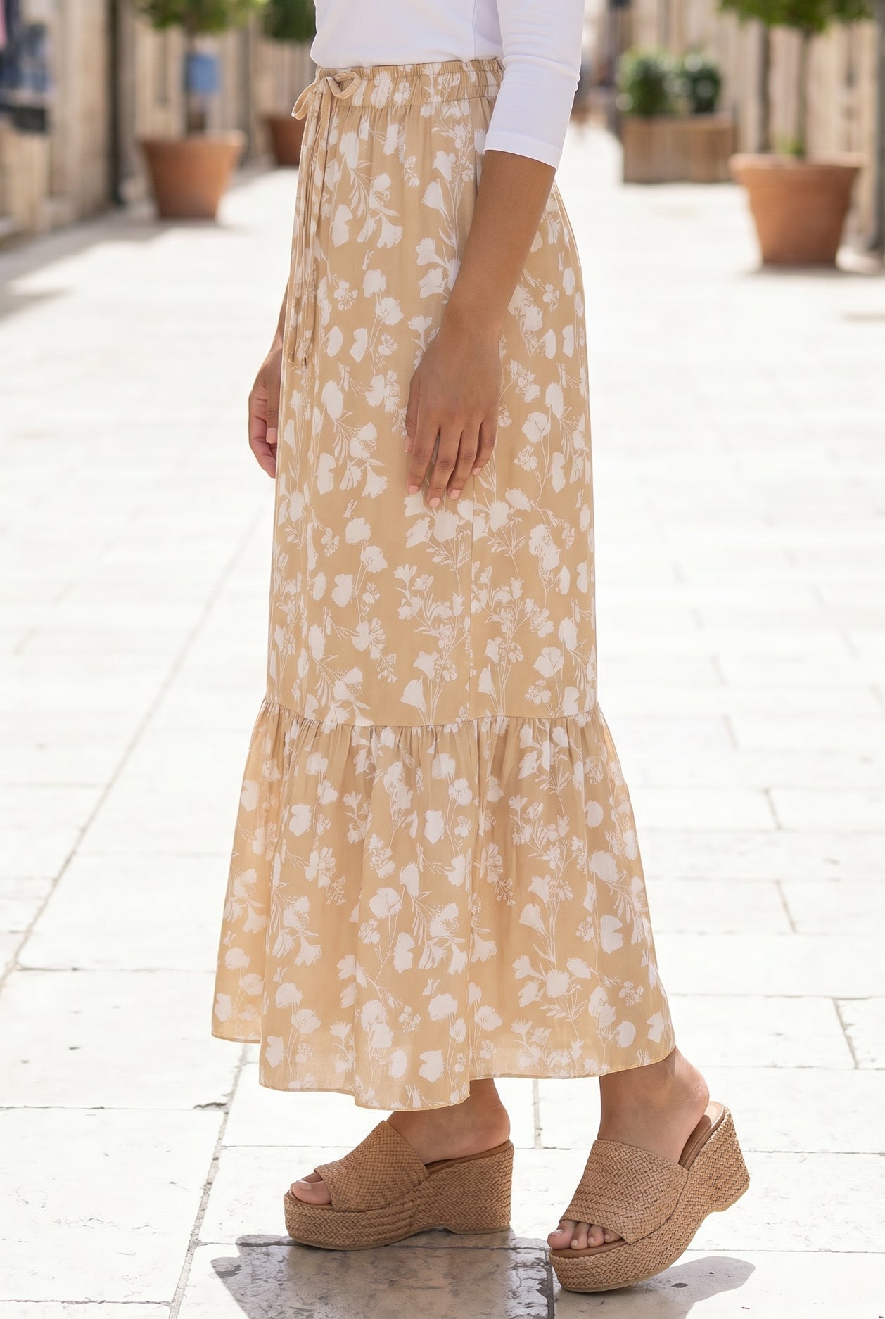 Woman in a white top and yellow floral skirt standing on a stone-paved street.