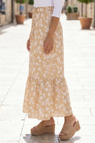Woman in a white top and yellow floral skirt standing on a stone-paved street.