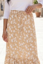 Woman in a white top and yellow floral skirt standing on a stone-paved street.