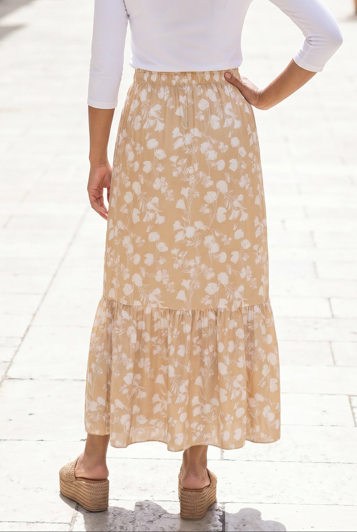 Woman in a white top and yellow floral skirt standing on a stone-paved street.