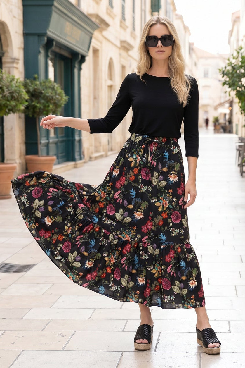 Woman in a black top and floral skirt standing on a street with potted plants and buildings.