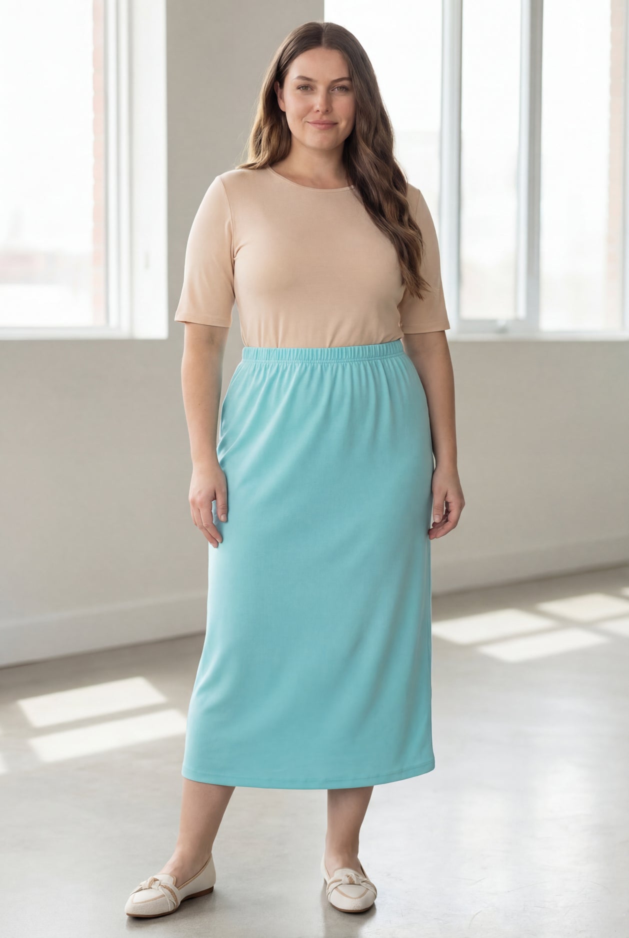 Woman wearing a beige shirt & light blue maxi length pencil skirt standing in a bright room with large windows.