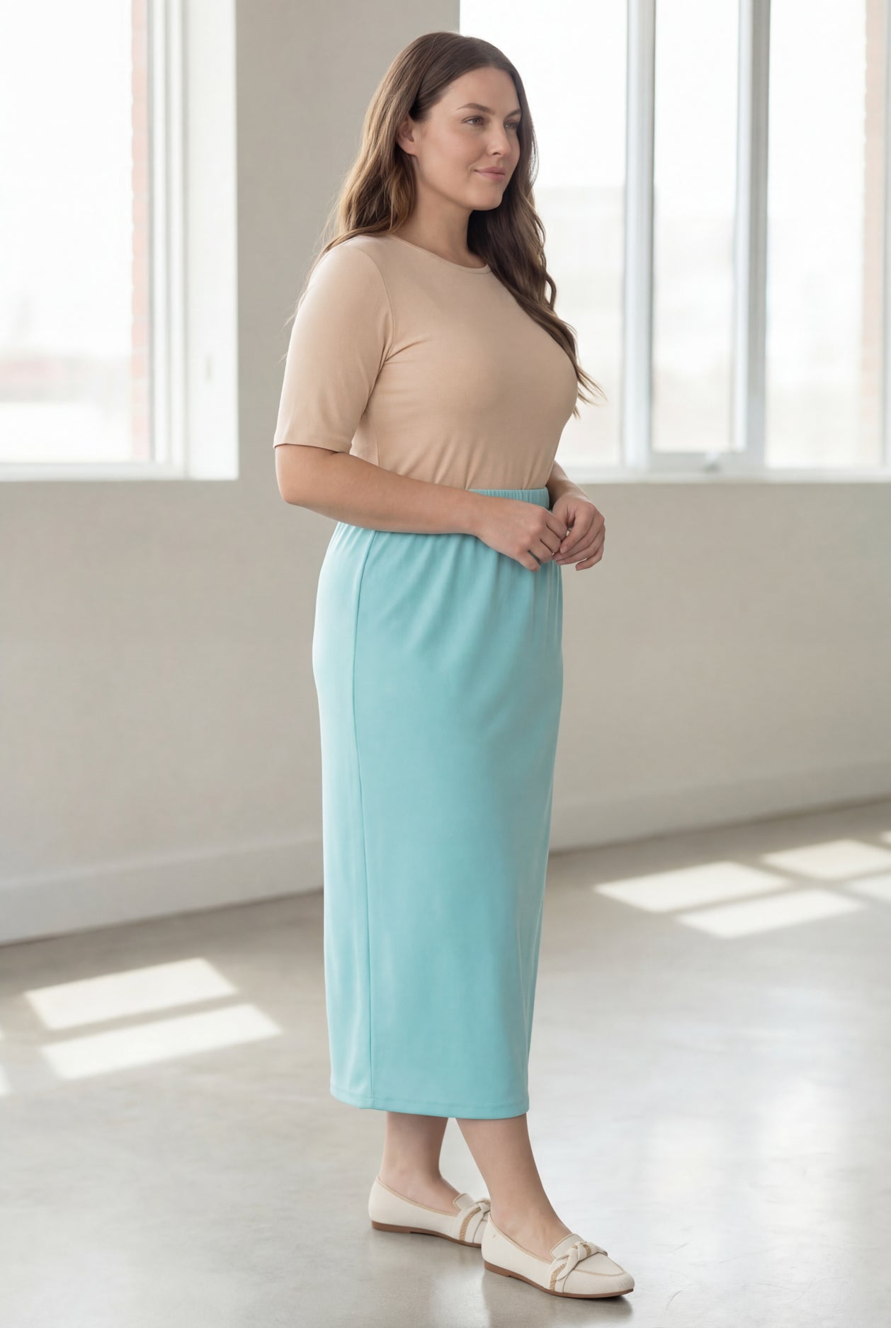 Woman wearing a beige shirt & light blue maxi length pencil skirt standing in a bright room with large windows.