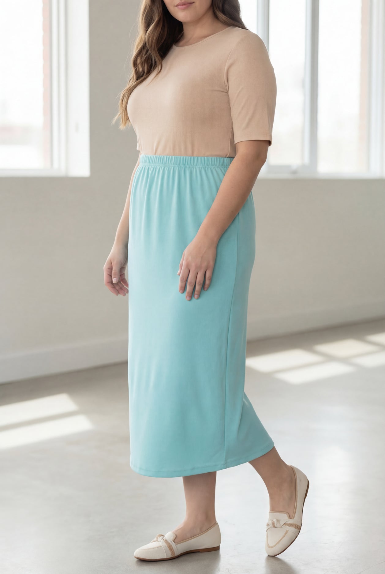 Woman wearing a beige shirt & light blue maxi length pencil skirt standing in a bright room with large windows.