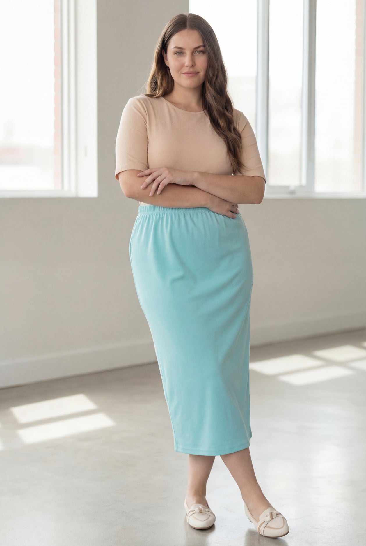 Woman wearing a beige shirt & light blue maxi length pencil skirt standing in a bright room with large windows.
