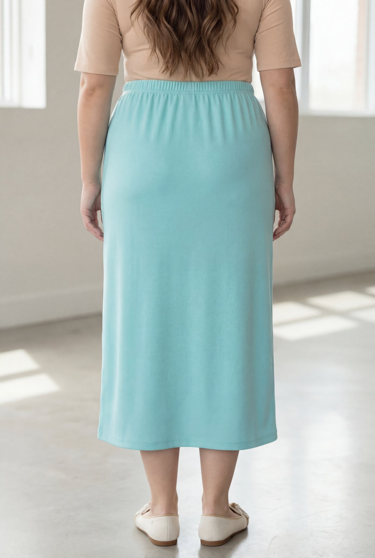 Woman wearing a beige shirt & light blue maxi length pencil skirt standing in a bright room with large windows.