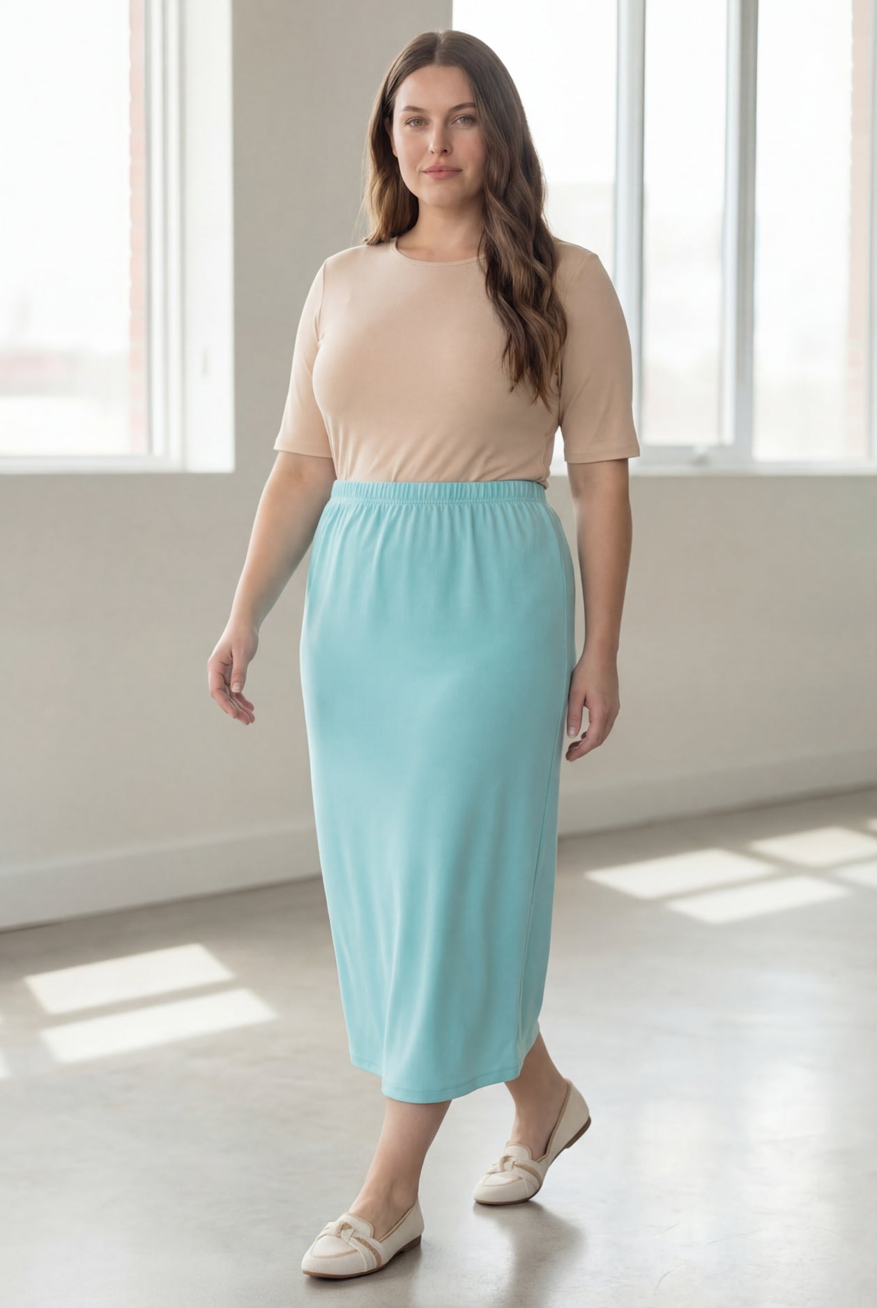 Woman wearing a beige shirt & light blue maxi length pencil skirt standing in a bright room with large windows.
