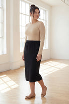 Woman wearing a beige top and  black skirt in a bright room.