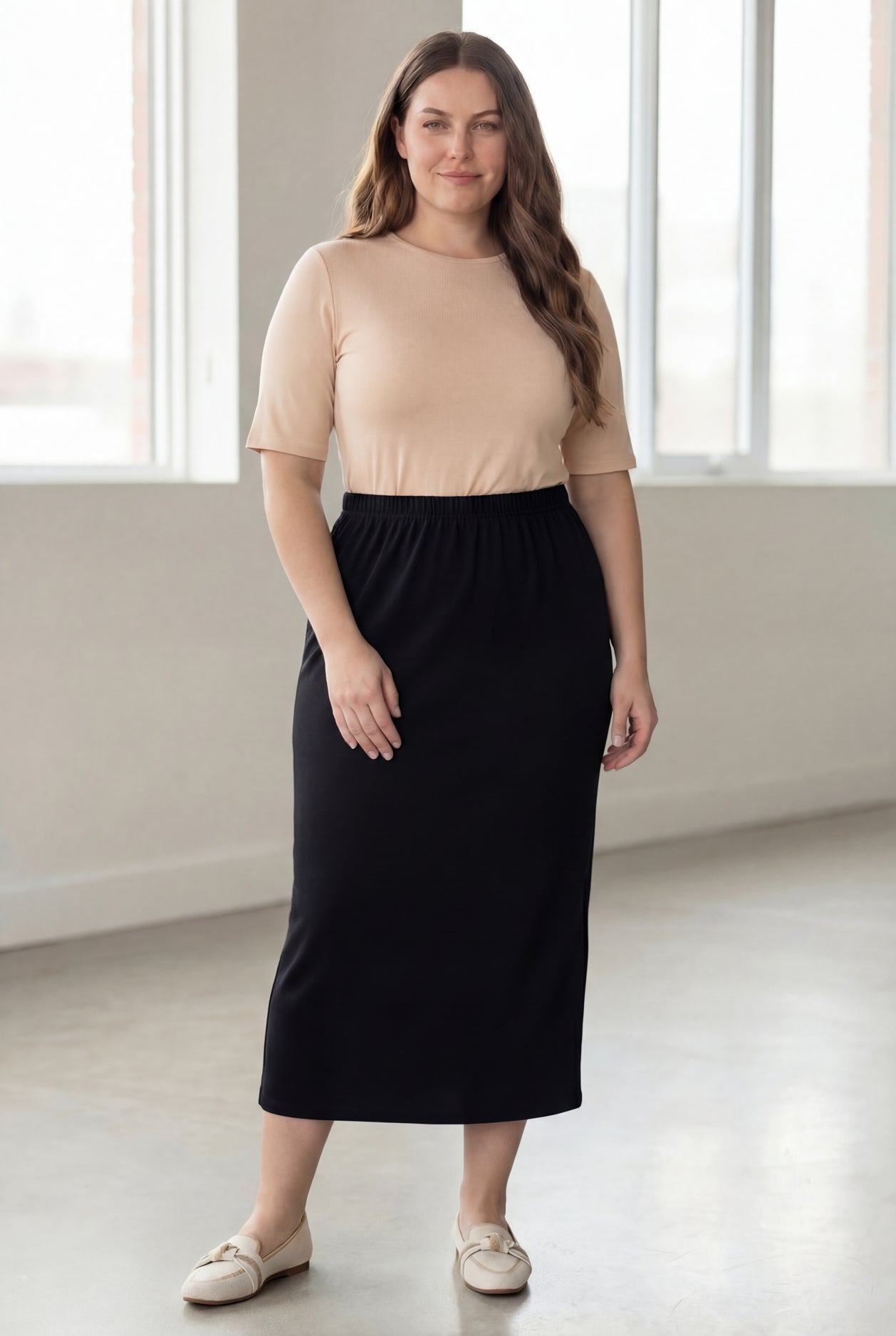 Woman wearing a beige shirt & black maxi length pencil skirt standing in a bright room with large windows.
