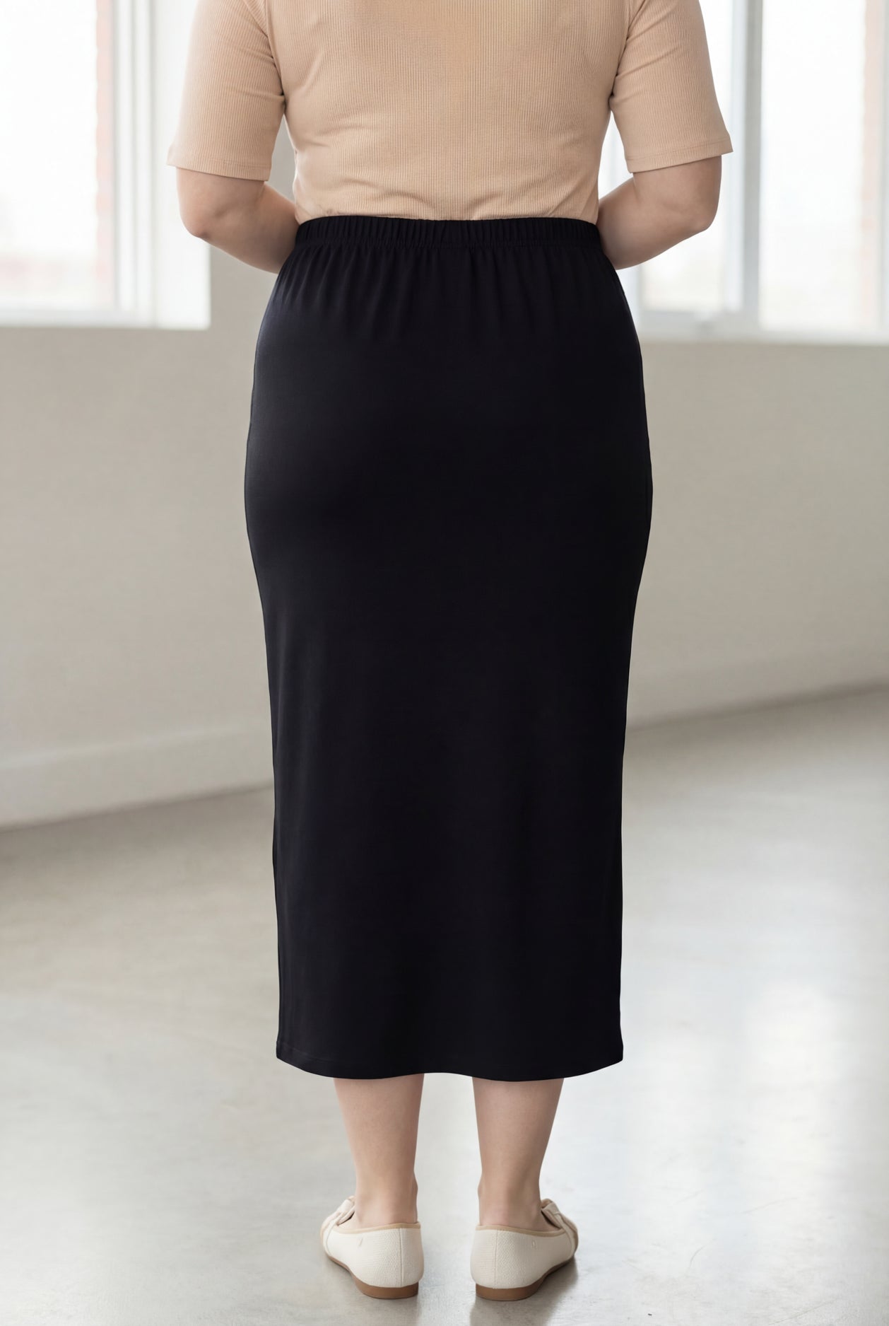 Woman wearing a beige shirt & black maxi length pencil skirt standing in a bright room with large windows.