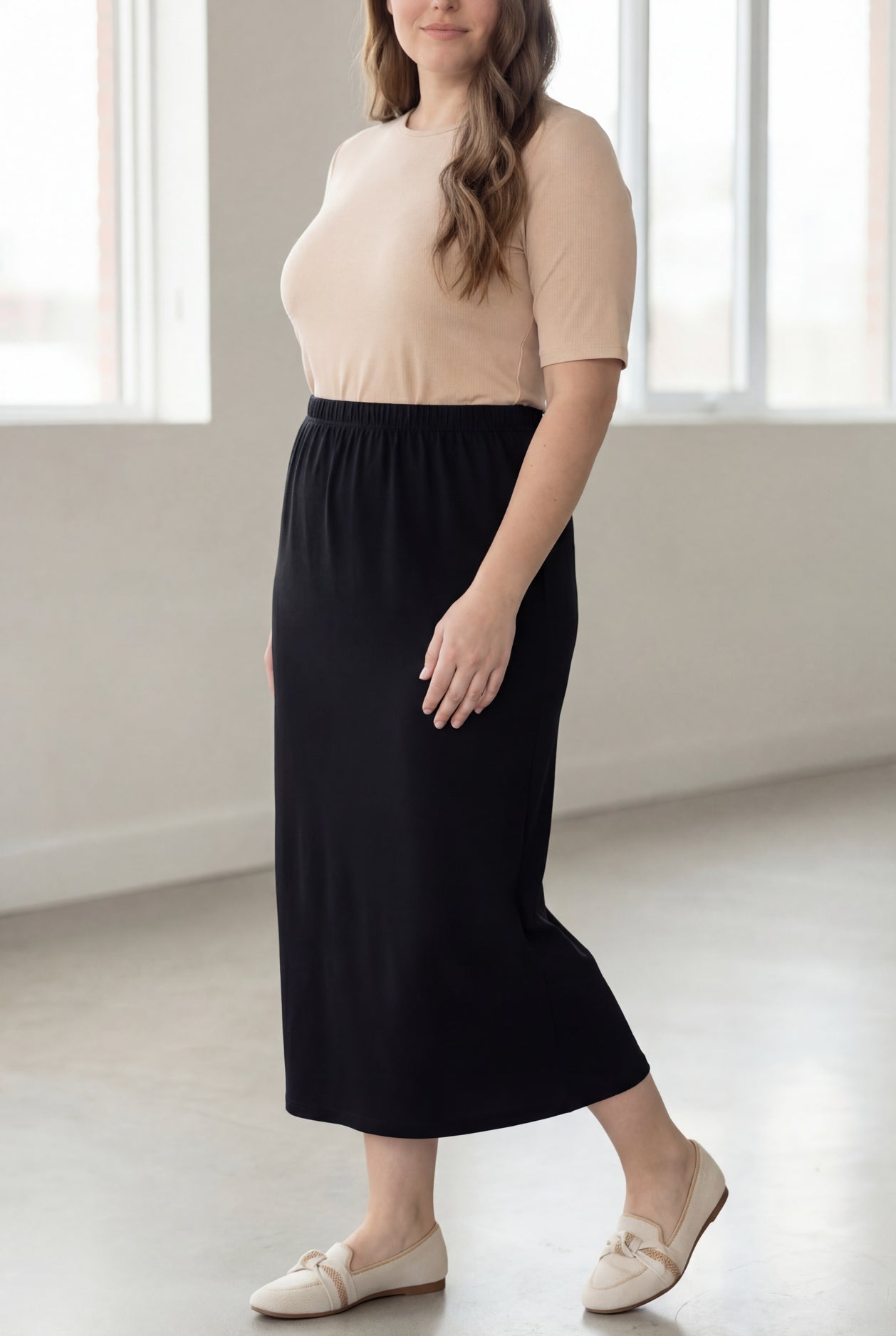 Woman wearing a beige shirt & black maxi length pencil skirt standing in a bright room with large windows.