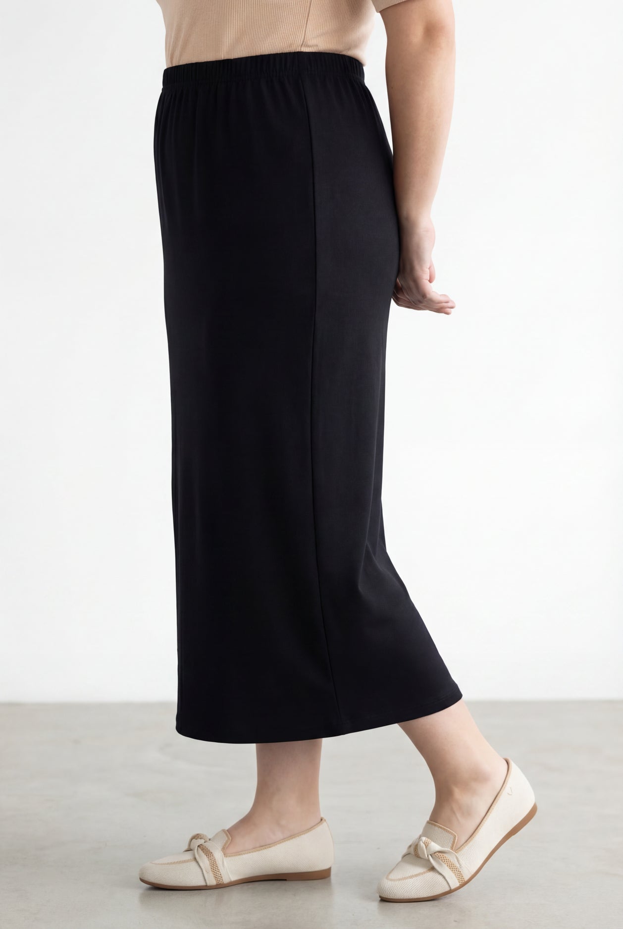 Woman wearing a beige shirt & black maxi length pencil skirt standing in a bright room with large windows.