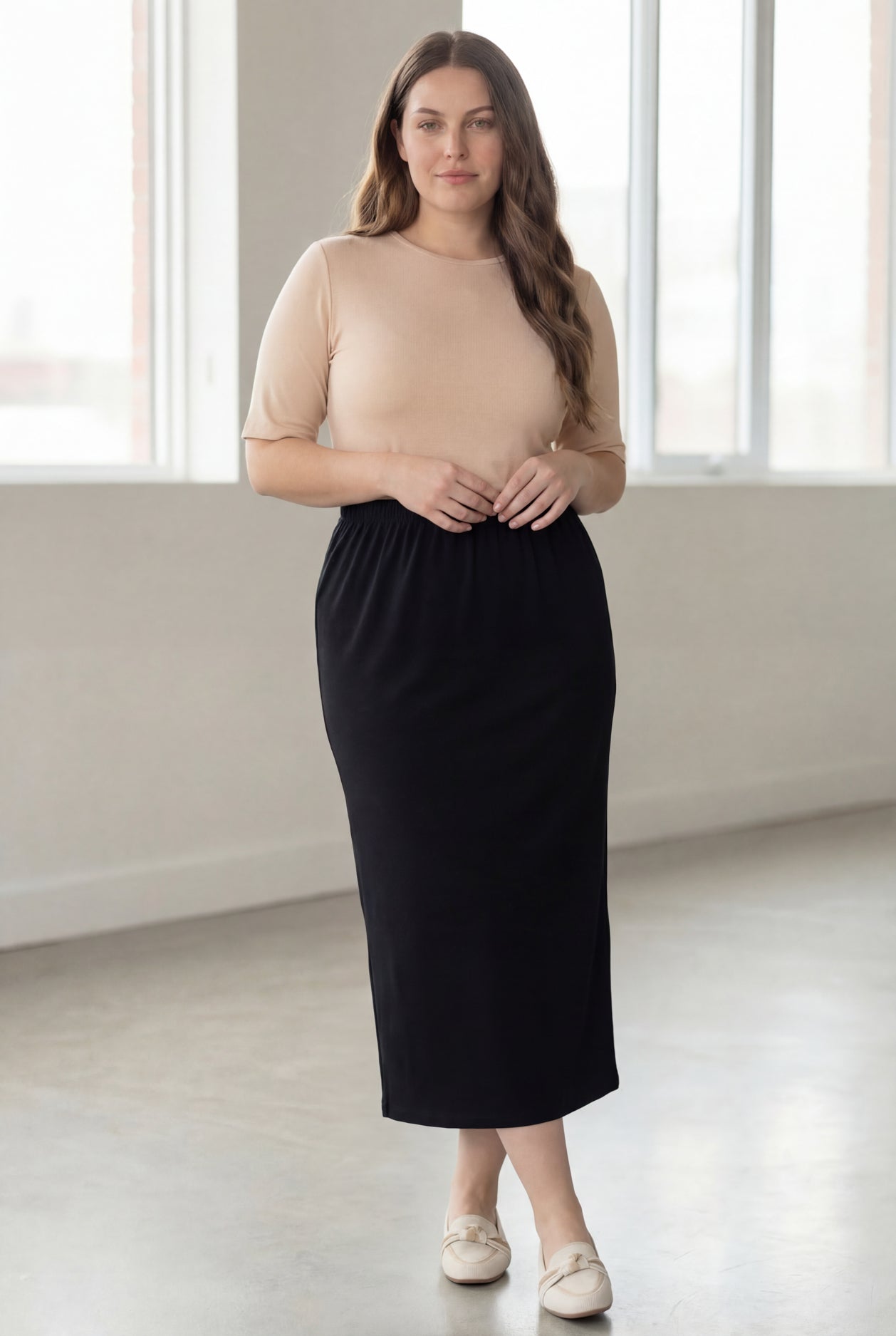 Woman wearing a beige shirt & black maxi length pencil skirt standing in a bright room with large windows.