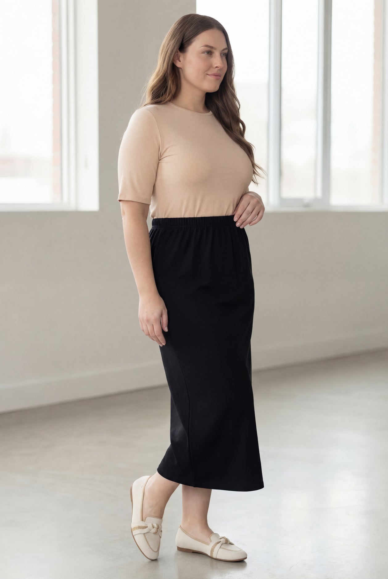 Woman wearing a beige shirt & black maxi length pencil skirt standing in a bright room with large windows.