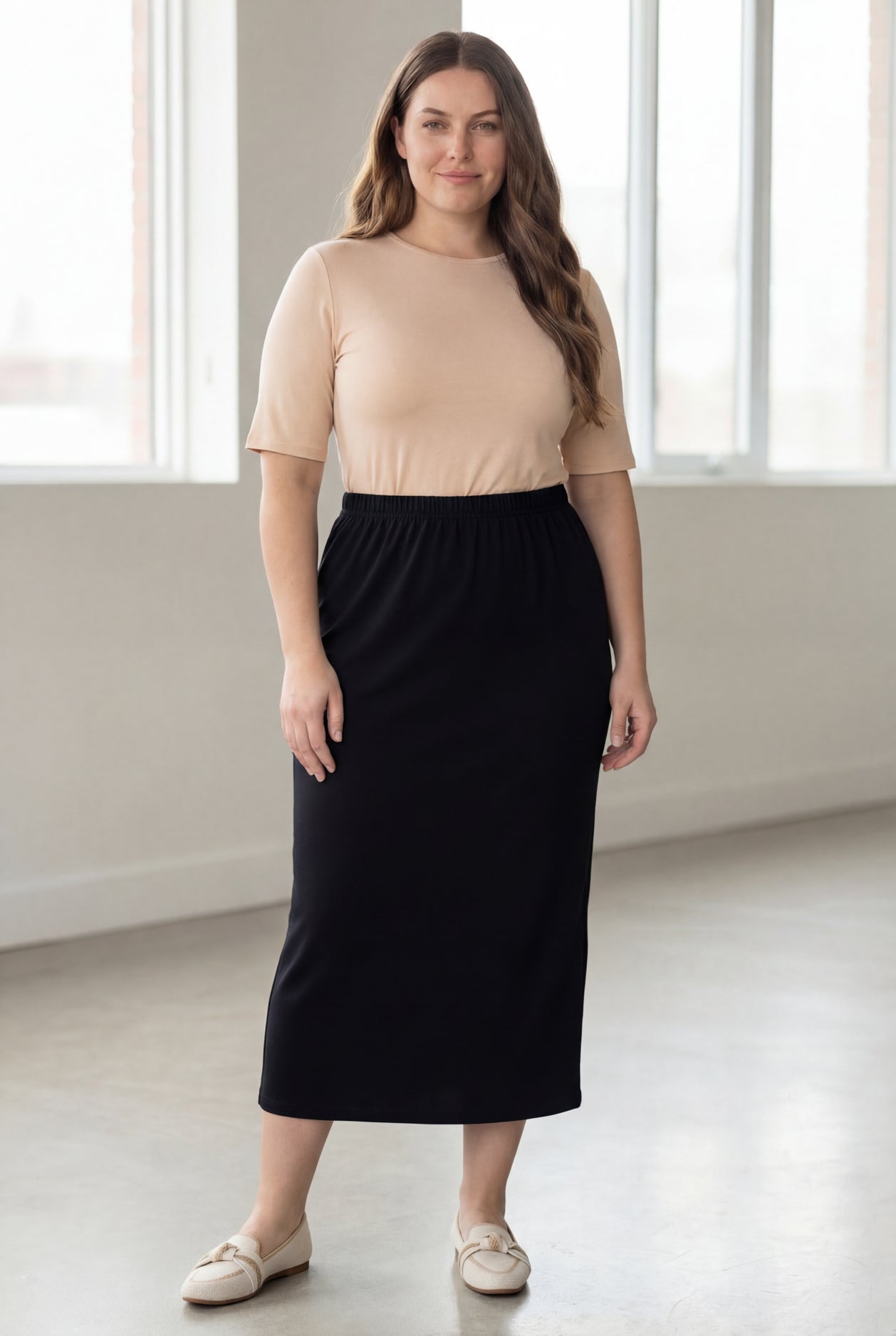 Woman wearing a beige shirt & black maxi length pencil skirt standing in a bright room with large windows.