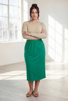 Woman wearing a beige top and green skirt in a bright room with large windows.