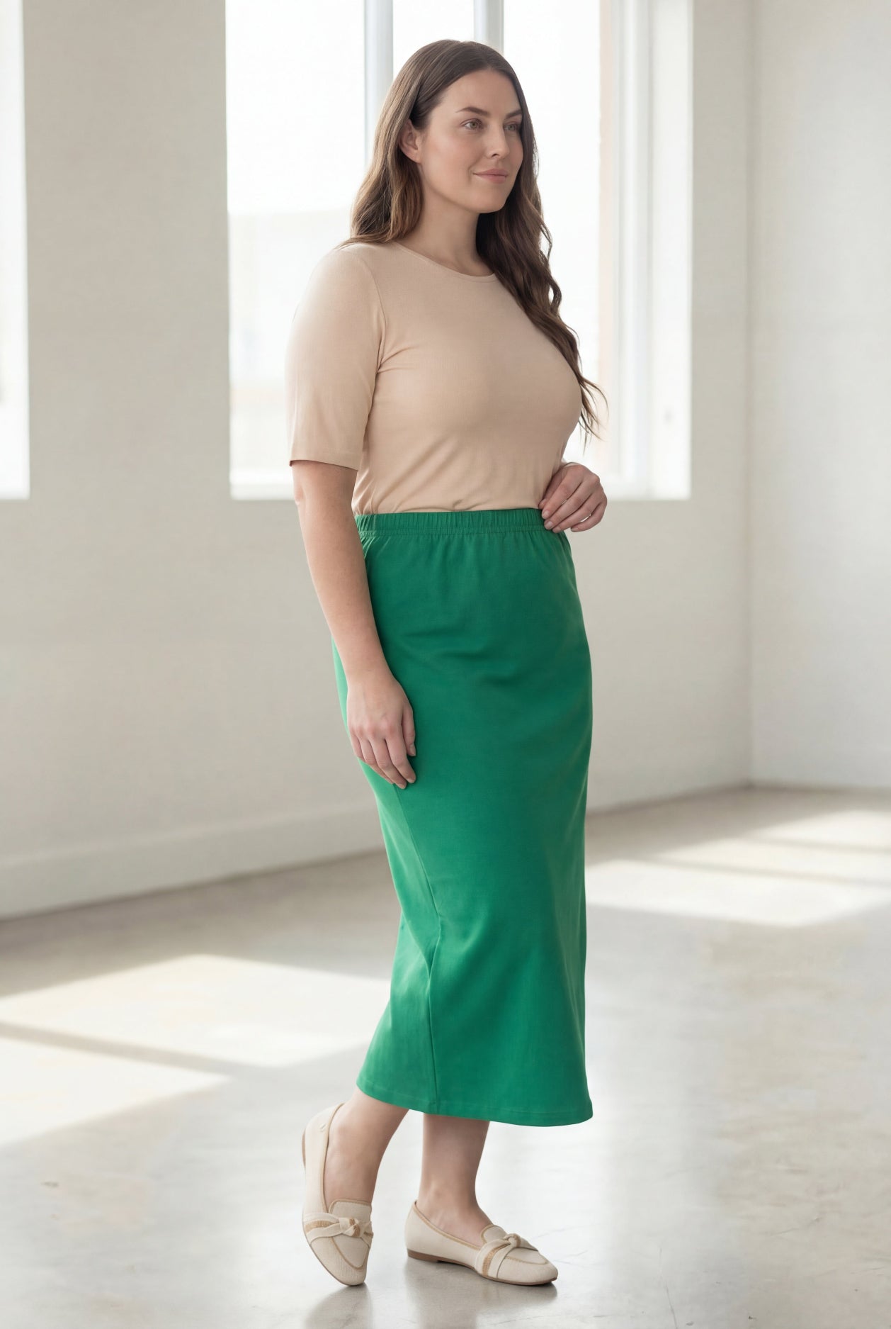 Woman wearing a beige shirt & green maxi length pencil skirt standing in a bright room with large windows.