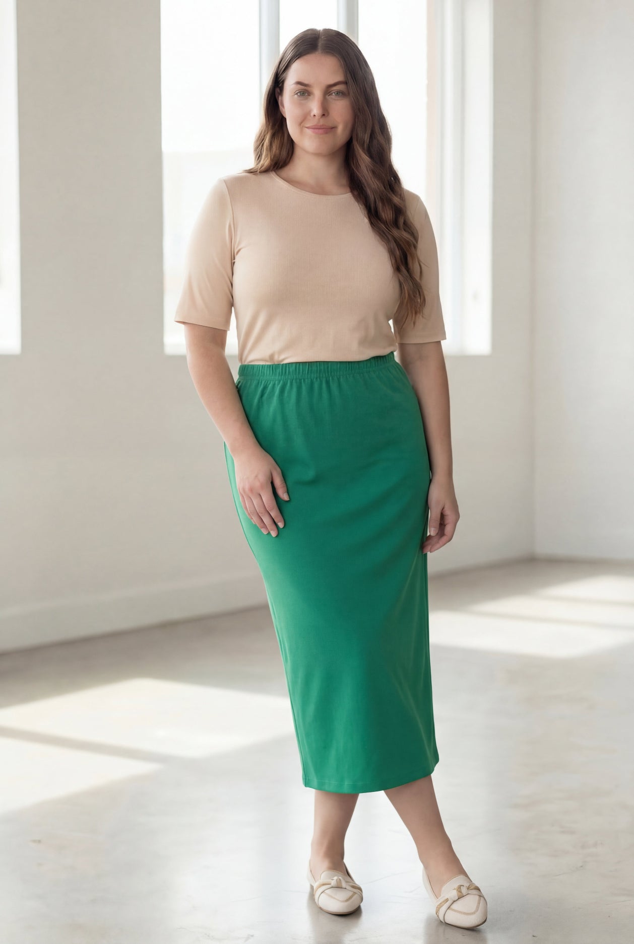 Woman wearing a beige shirt & green maxi length pencil skirt standing in a bright room with large windows.