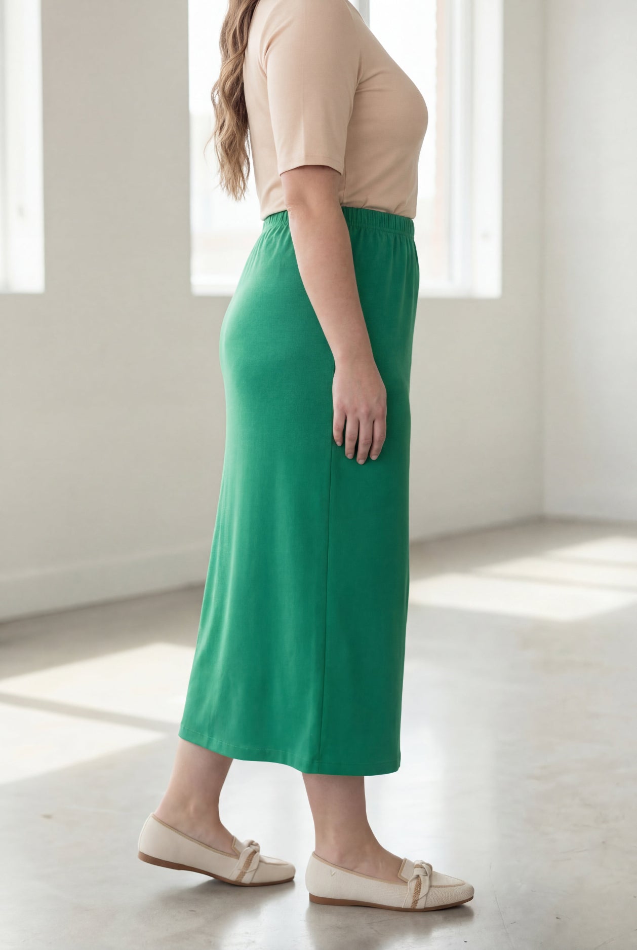 Woman wearing a beige shirt & green maxi length pencil skirt standing in a bright room with large windows.