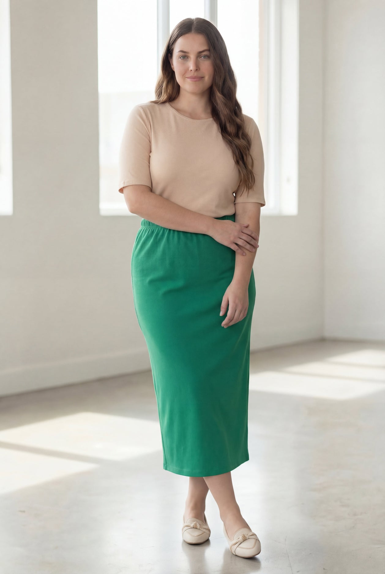 Woman wearing a beige shirt & green maxi length pencil skirt standing in a bright room with large windows.