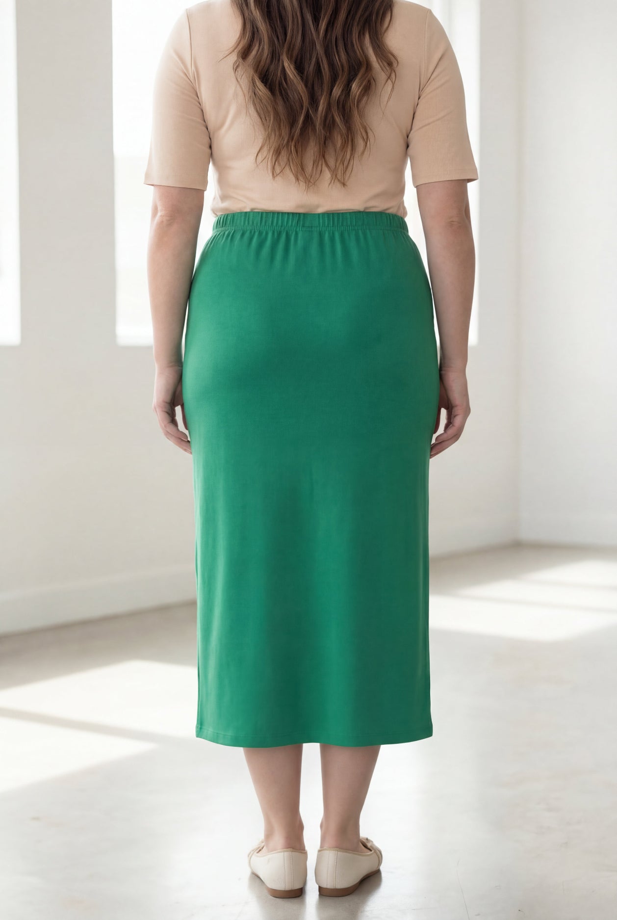 Woman wearing a beige shirt & green maxi length pencil skirt standing in a bright room with large windows.