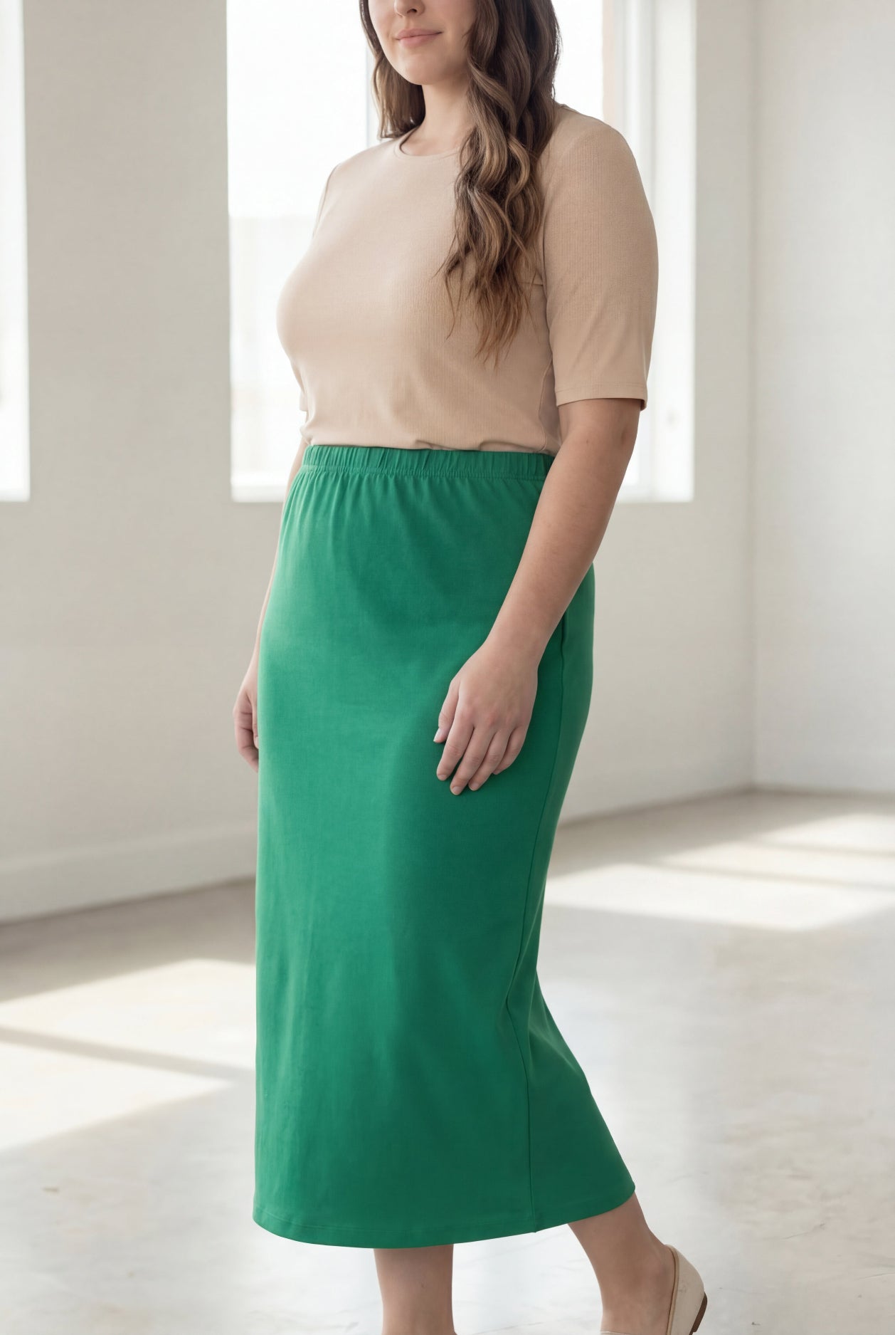 Woman wearing a beige shirt & green maxi length pencil skirt standing in a bright room with large windows.