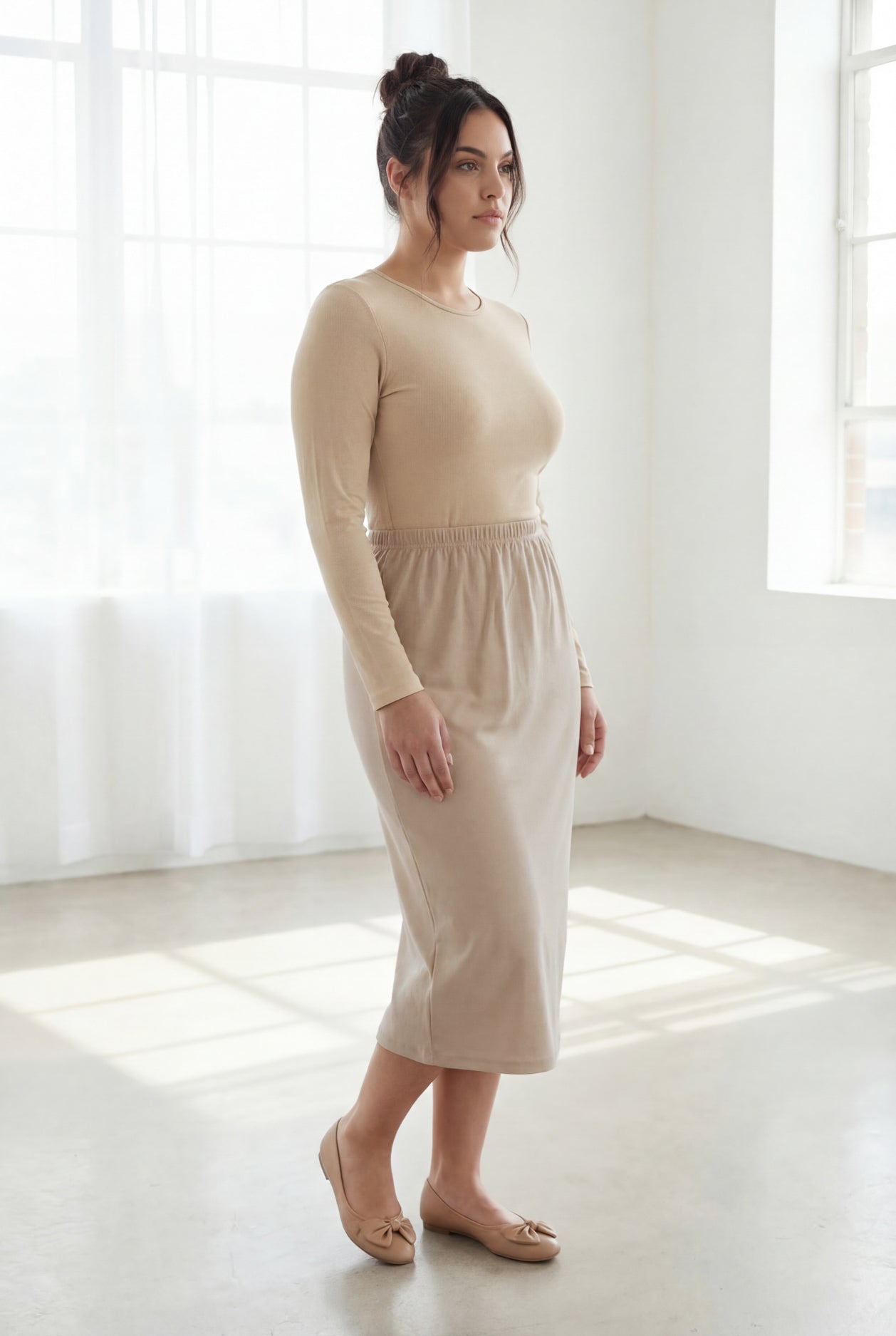 Woman wearing a beige top and beige skirt in a bright room with large windows.
