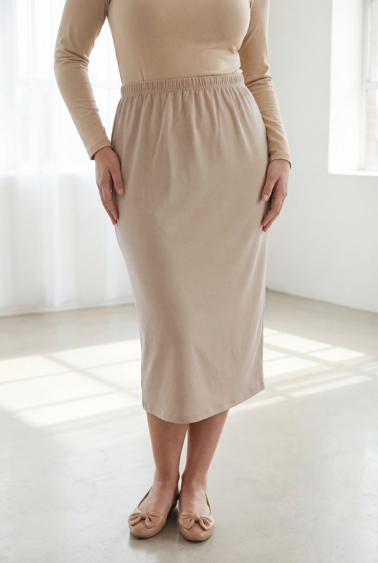 Woman wearing a beige top and beige skirt in a bright room with large windows.