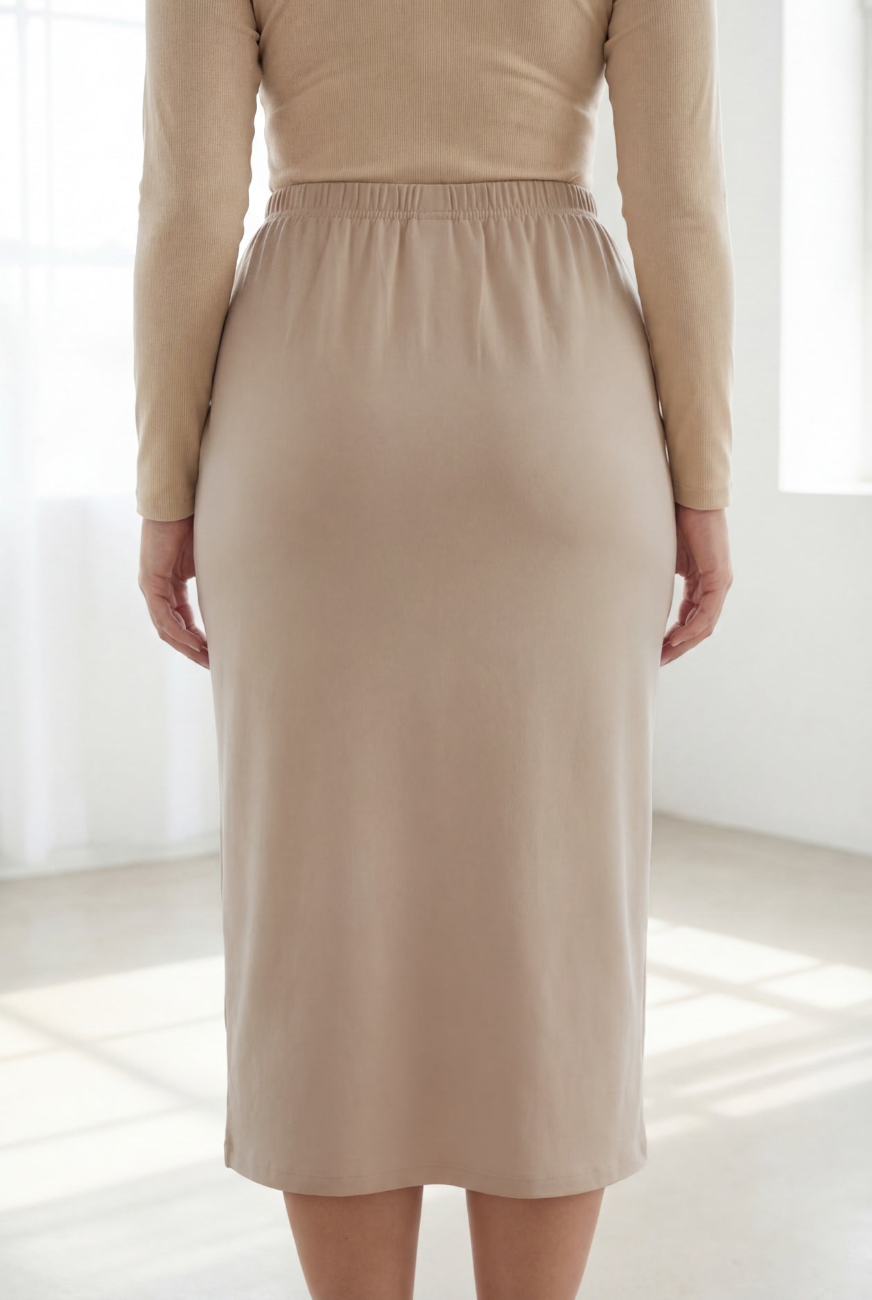Woman wearing a beige top and beige skirt in a bright room with large windows.