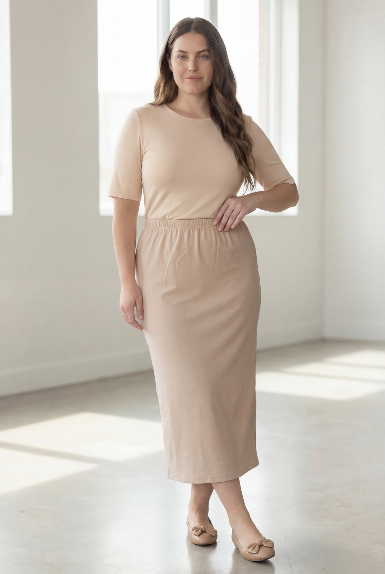 Woman wearing a beige shirt & beige maxi length pencil skirt standing in a bright room with large windows.
