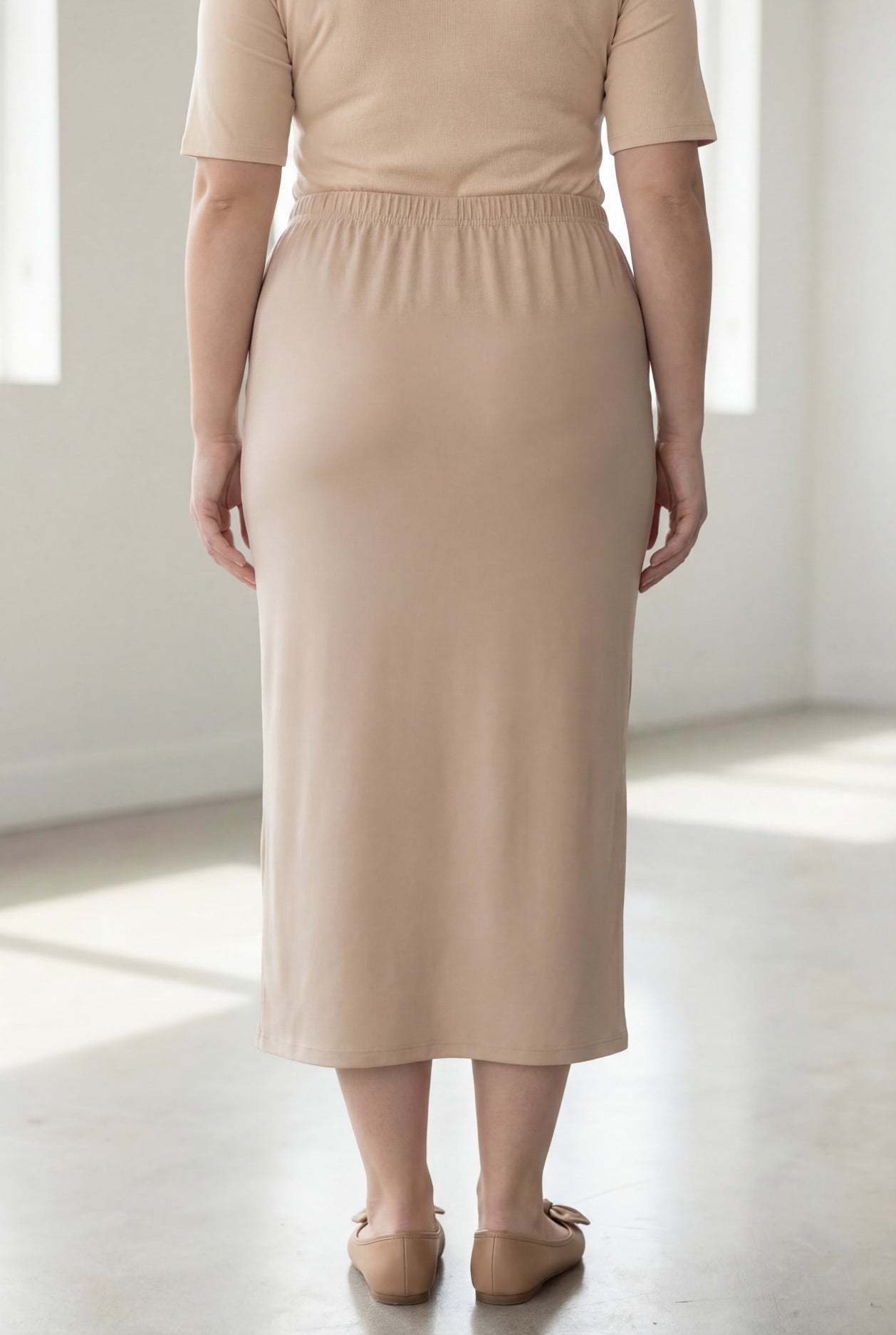 Woman wearing a beige shirt & beige maxi length pencil skirt standing in a bright room with large windows.