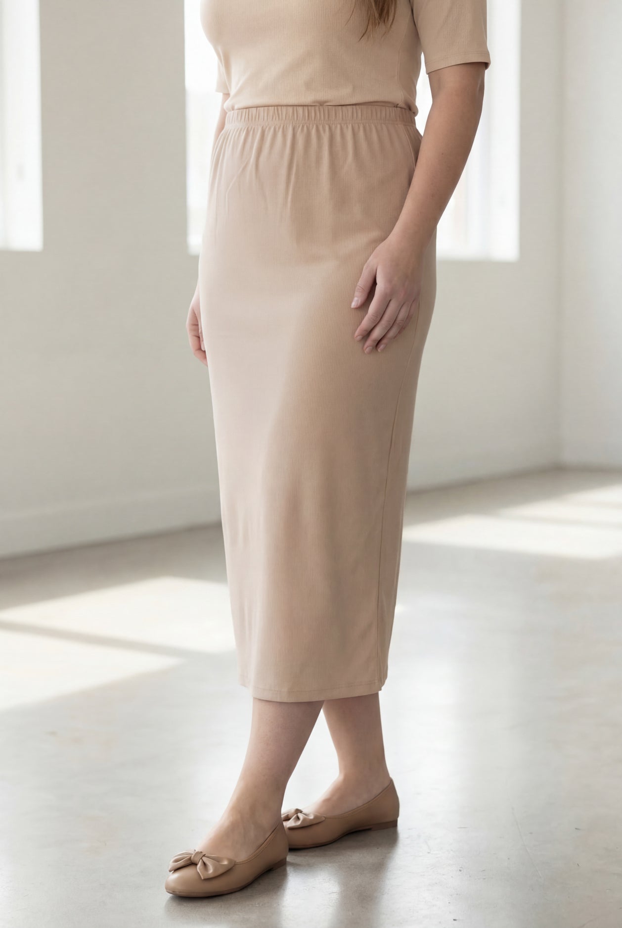 Woman wearing a beige shirt & beige maxi length pencil skirt standing in a bright room with large windows.