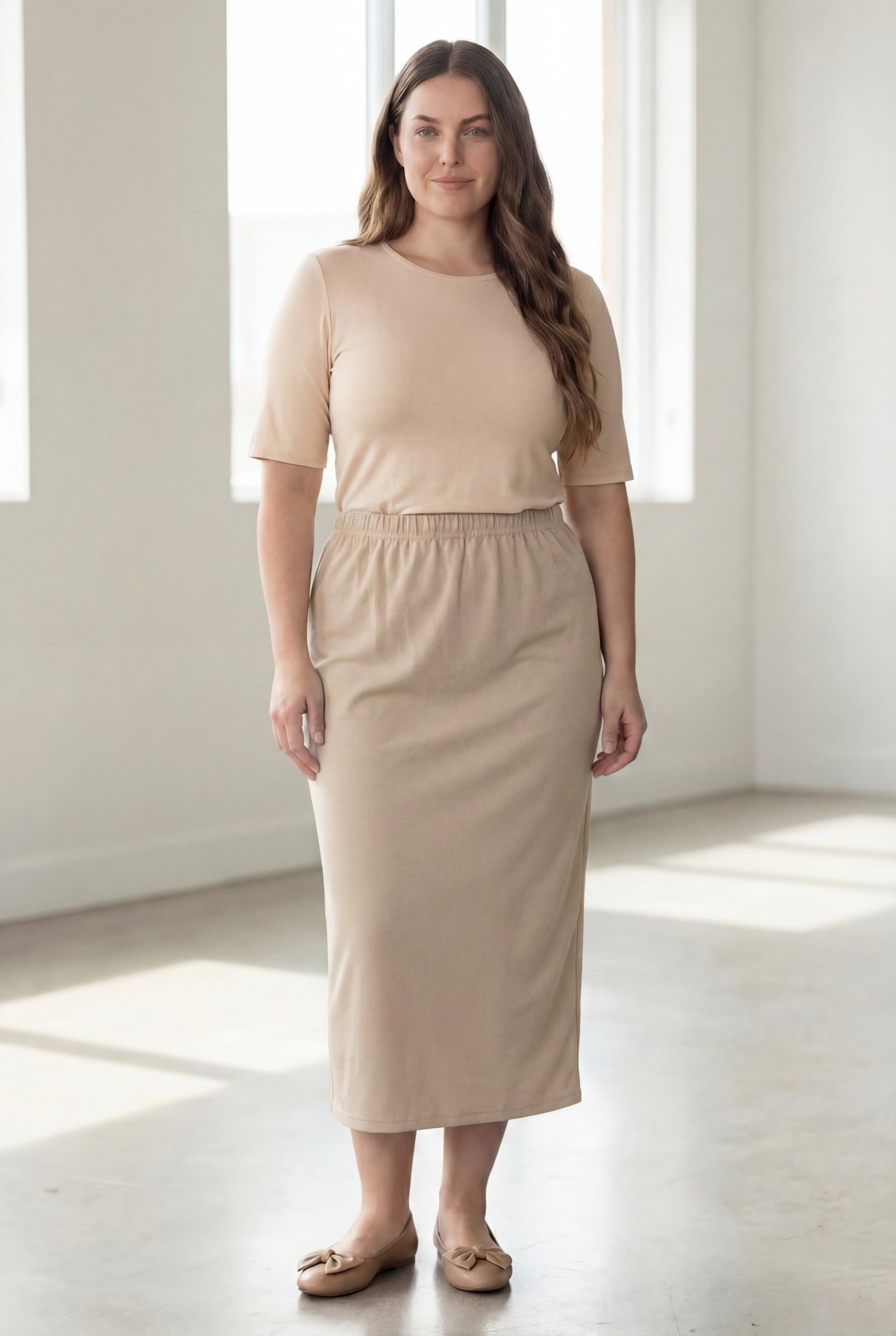 Woman wearing a beige shirt & beige maxi length pencil skirt standing in a bright room with large windows.