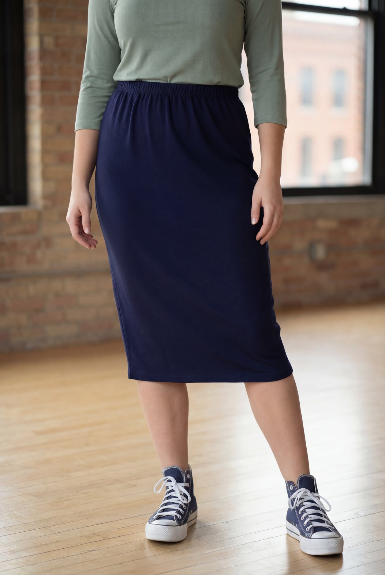 Person wearing a navy 27" Classic skirt and green top in an indoor setting with brick walls.