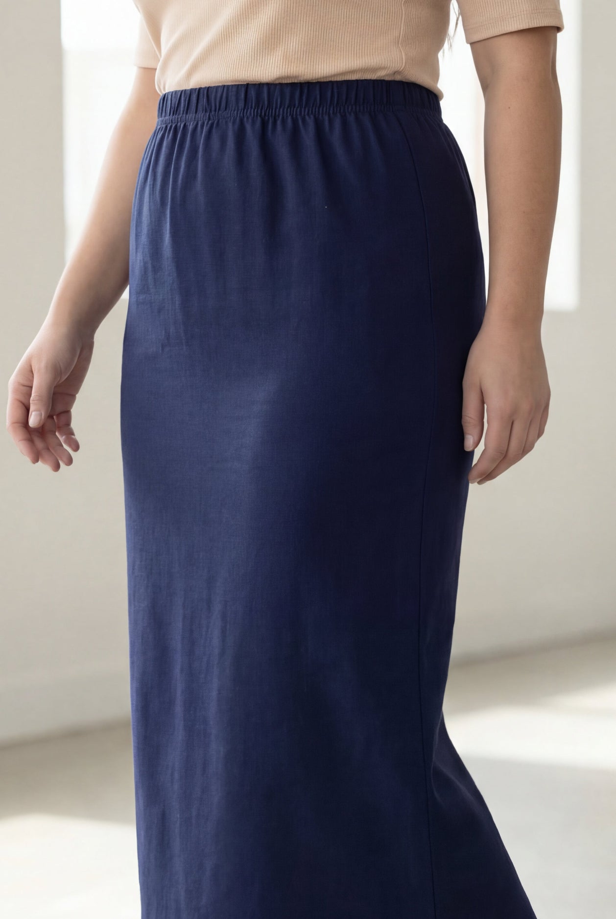 Woman wearing a beige shirt & blue maxi length pencil skirt standing in a bright room with large windows.