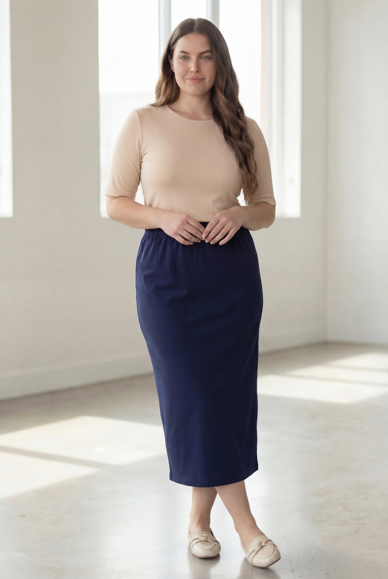 Woman wearing a beige shirt & blue maxi length pencil skirt standing in a bright room with large windows.
