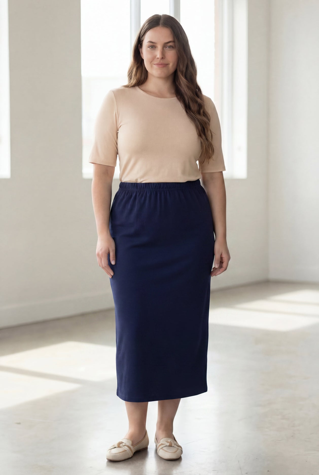 Woman wearing a beige shirt & blue maxi length pencil skirt standing in a bright room with large windows.
