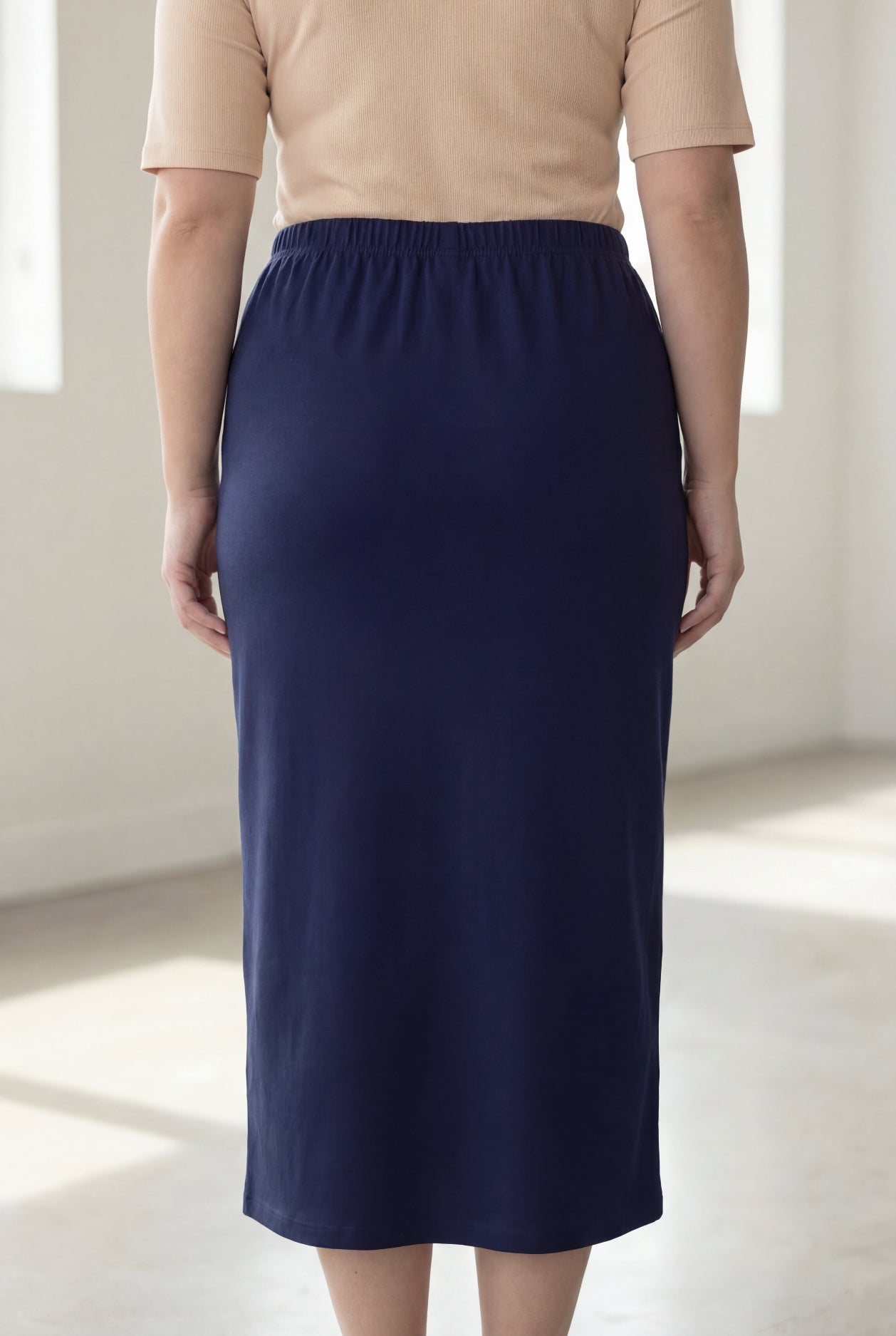 Woman wearing a beige shirt & blue maxi length pencil skirt standing in a bright room with large windows.