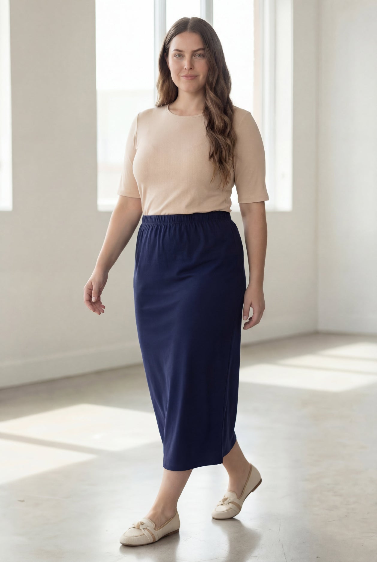 Woman wearing a beige shirt & blue maxi length pencil skirt standing in a bright room with large windows.