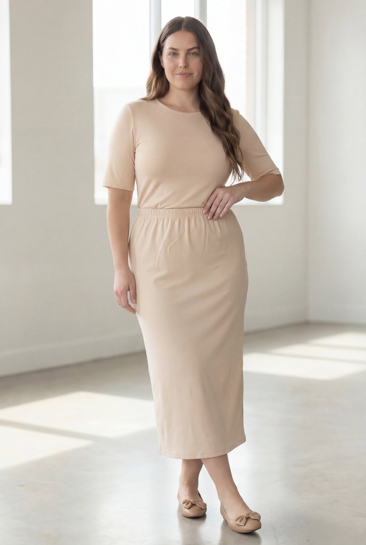 Woman wearing a beige shirt & maxi length pencil skirt standing in a bright room with large windows.