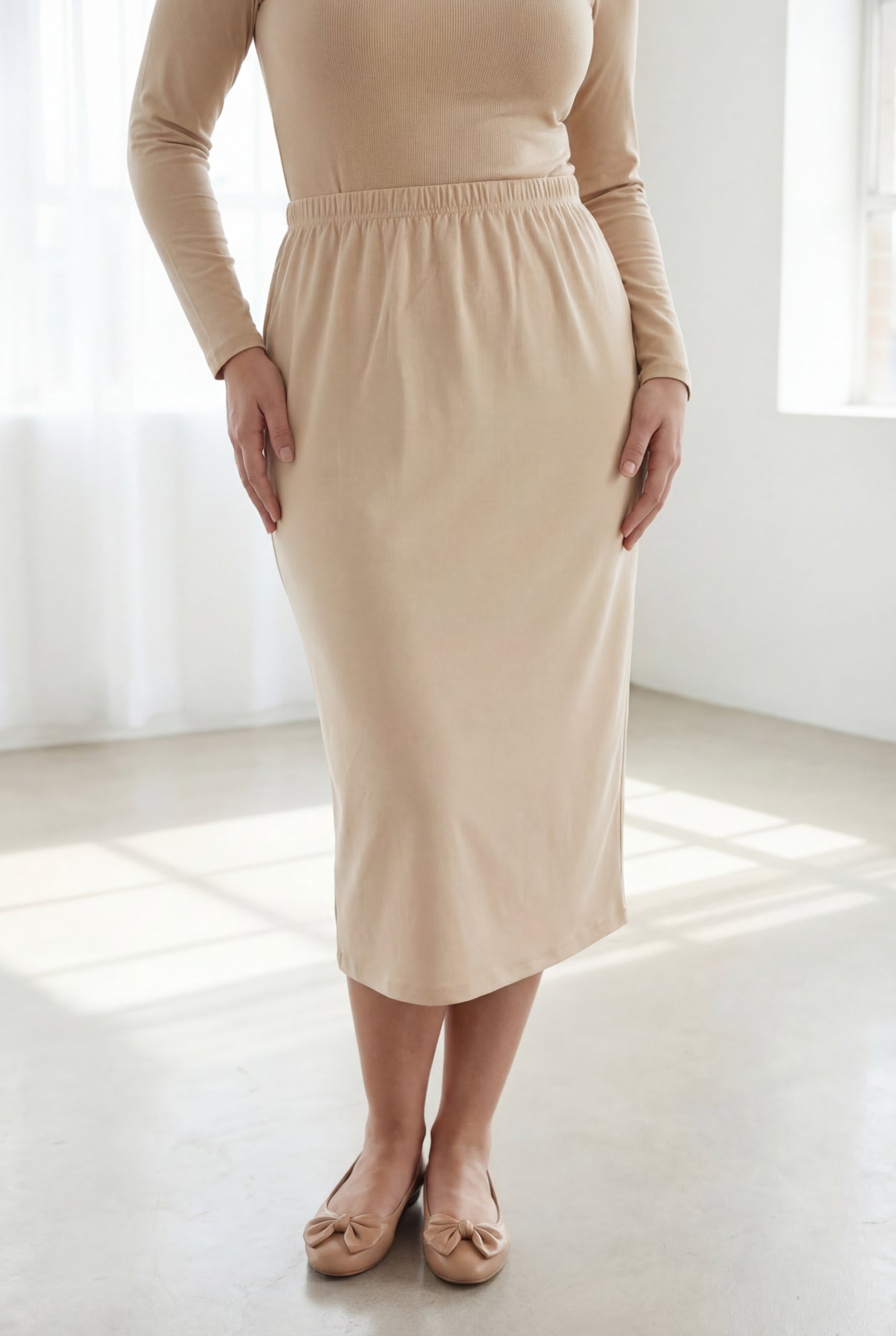 Woman wearing a beige shirt & skirt standing in a bright, minimalistic room.