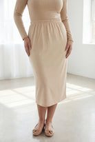 Woman wearing a beige shirt & skirt standing in a bright, minimalistic room.