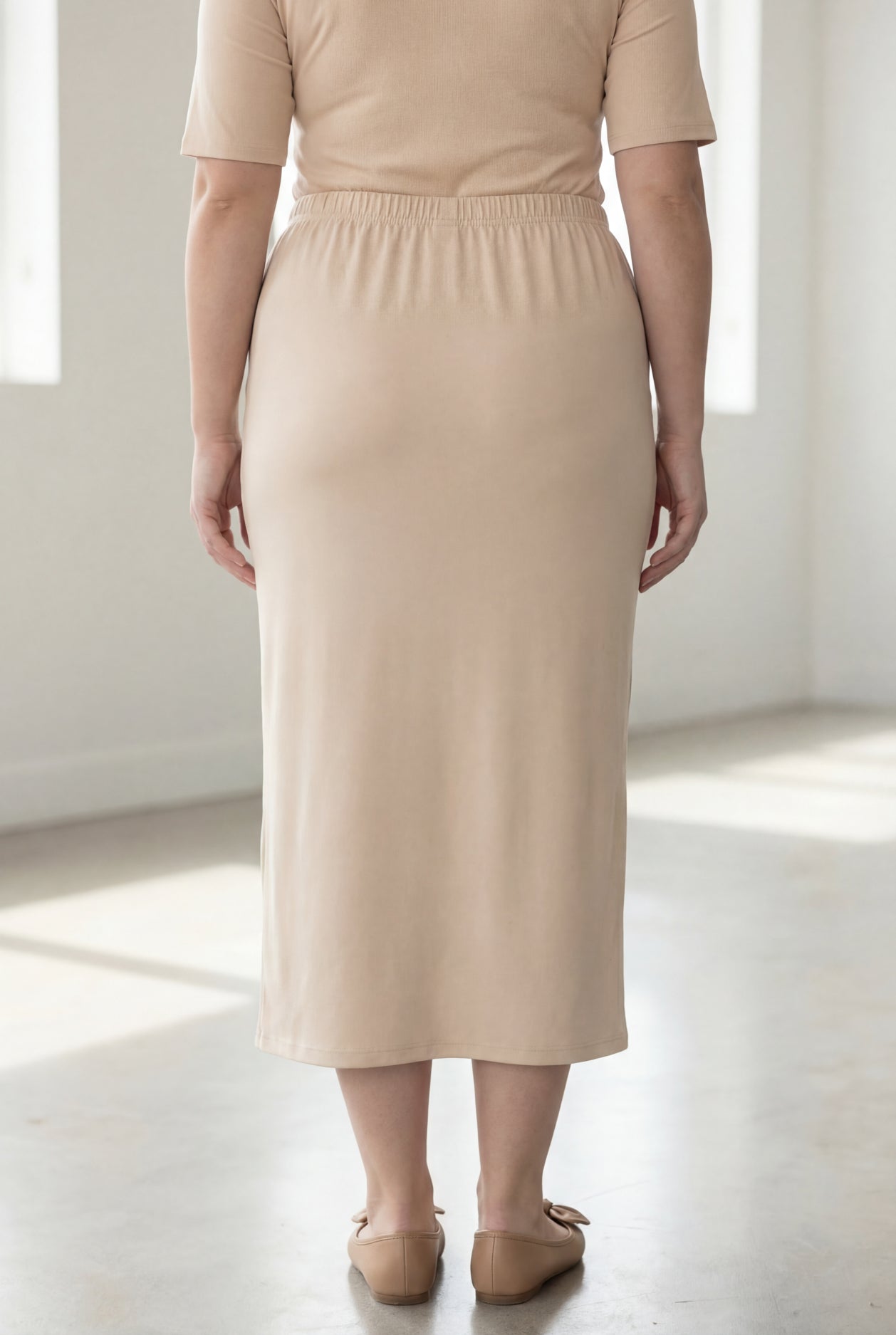 Woman wearing a beige shirt & maxi length pencil skirt standing in a bright room with large windows.